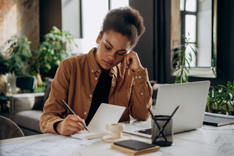 A dedicated woman with curly hair writing in a notebook at a modern office desk.
