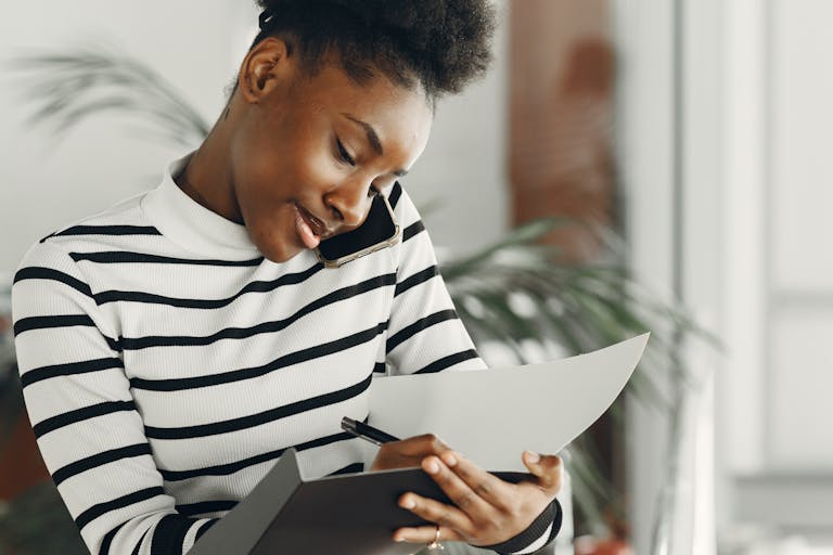 Young African woman multitasking with a phone call and taking notes indoors, wearing stripes.